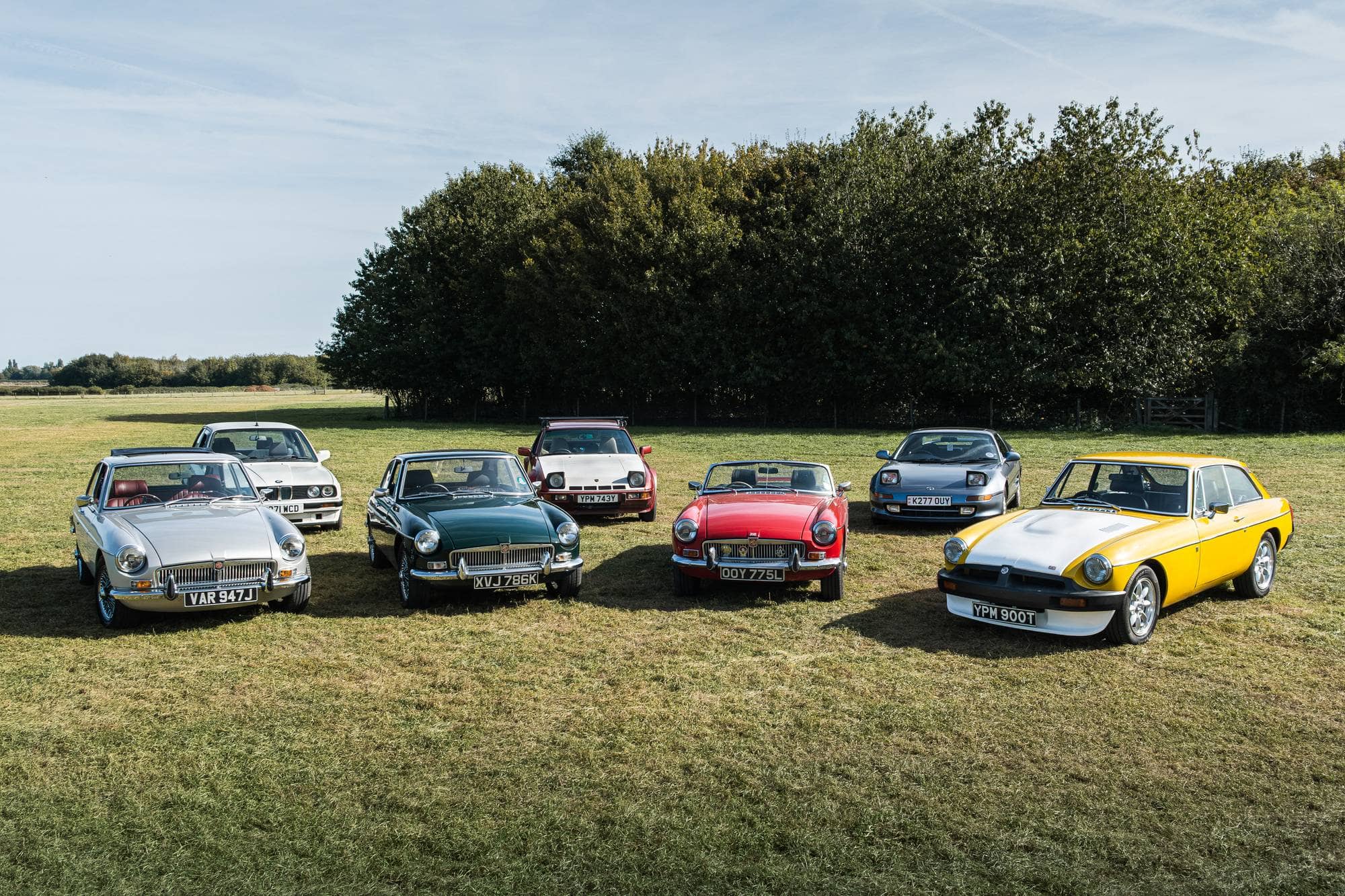 A lineup of classic MGs alongside a BMW E30, Porsche 924, and Toyota MR2 parked in a field