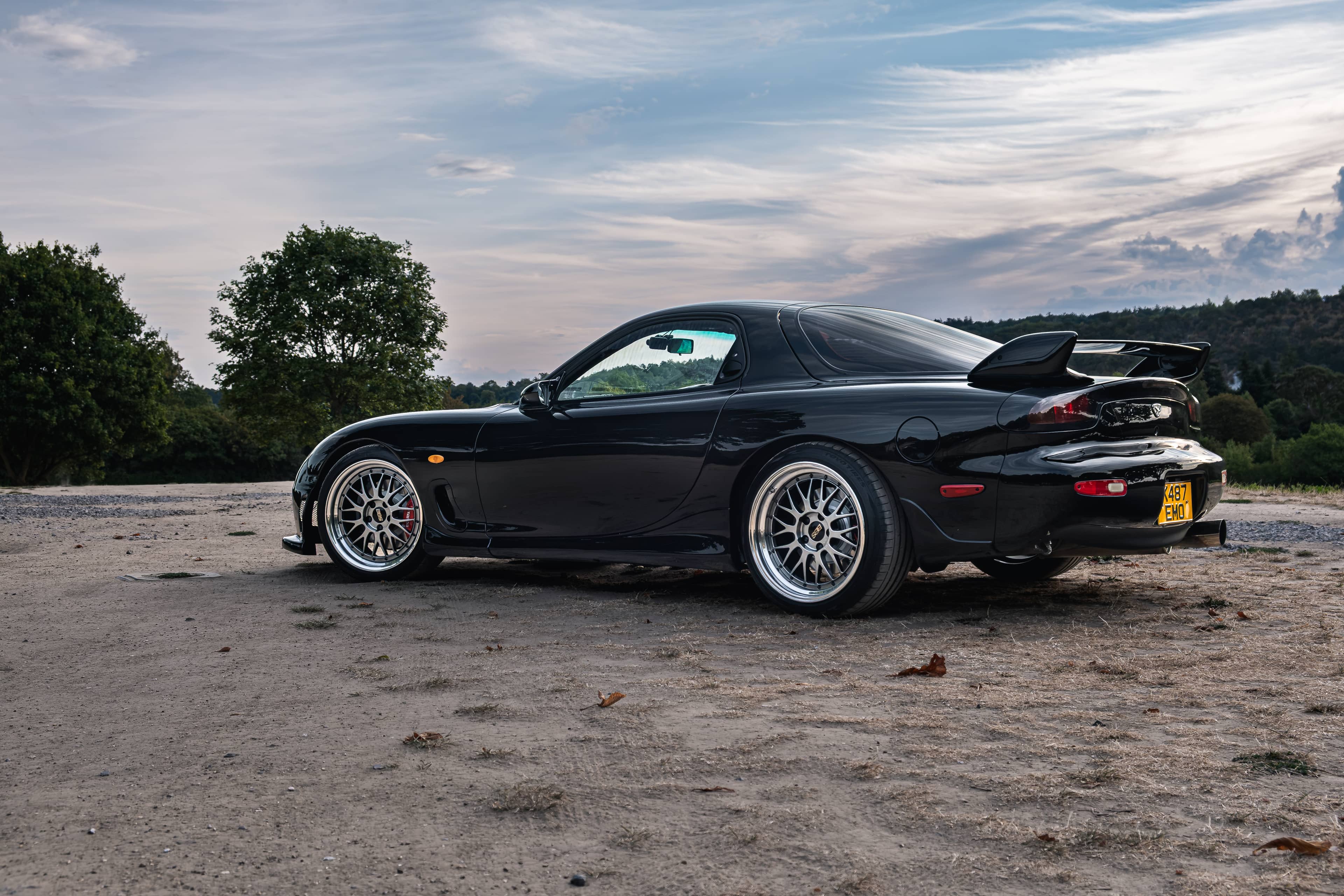 Black Mazda RX-7 FD3S on bronze BBS LM wheels, rear three-quarter view on a dirt car park at dawn with rolling hills behind